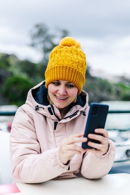 Mujer en ropa de invierno, sonriendo, mirando su teléfono celular, sentada en una terraza al aire libre