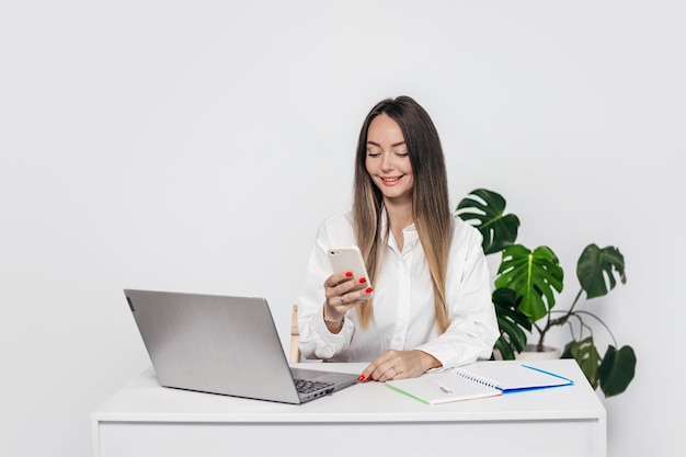 mujer con ropa de negocios se sienta en una mesa con una computadora portátil y mira el teléfono