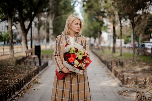 Mujer rubia en abrigo a cuadros con un brillante ramo de flores de pie en el callejón