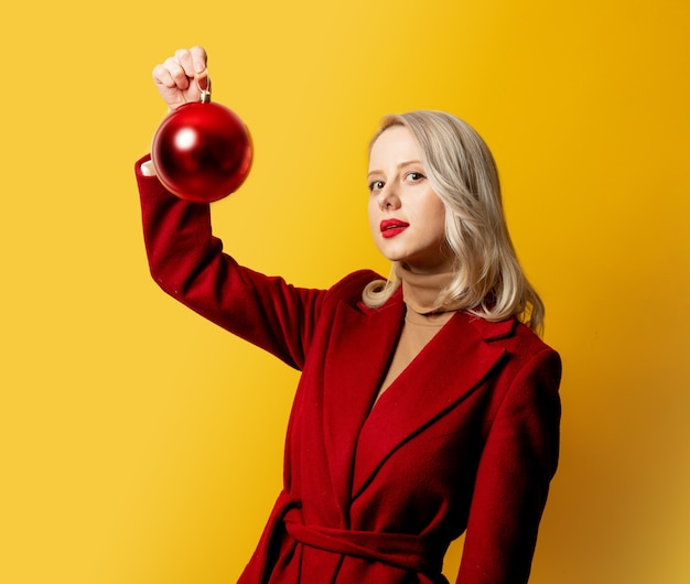 Mujer rubia en abrigo rojo con adorno de Navidad en pared amarilla