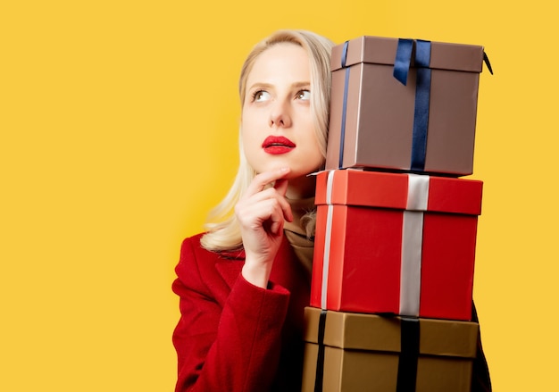 Mujer rubia en abrigo rojo con cajas de regalo en pared amarilla