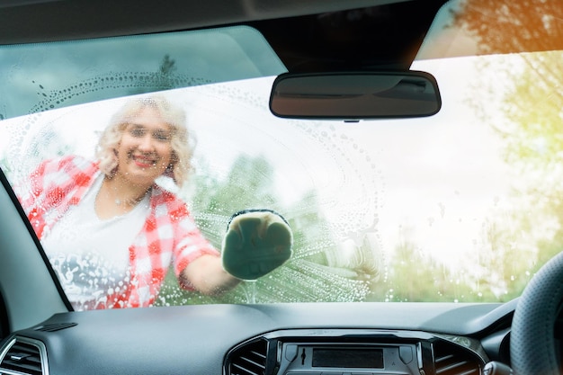 Una mujer rubia adulta lava el parabrisas de un auto con una toallita una vista desde el interior del auto