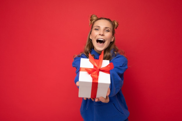 mujer rubia aislada sobre fondo rojo pared vistiendo sudadera con capucha de moda azul con caja de regalo y mirando a cámara y gritando.