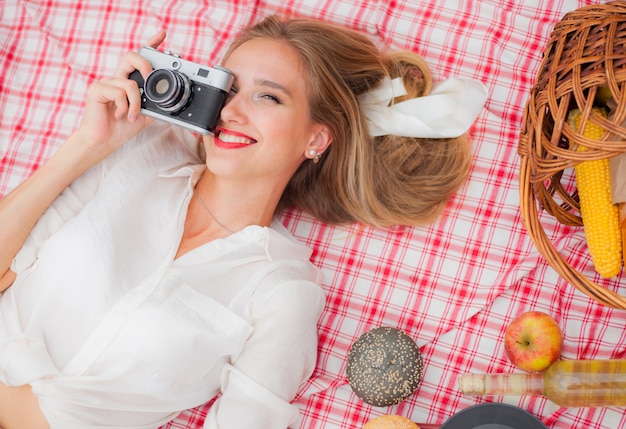 Mujer rubia alegre joven en la ropa del vintage que sostiene la cámara retra y que toma la foto mientras que miente en mantel de la comida campestre al aire libre. Vista superior