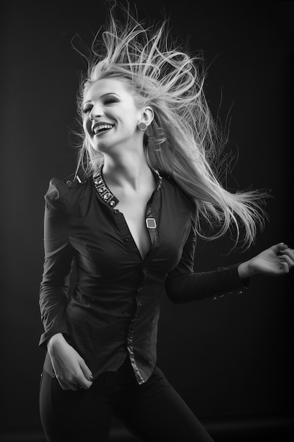 Mujer rubia alegre con maquillaje perfecto y cabello largo revoloteando viste blusa, posando en el estudio. Disparo en blanco y negro