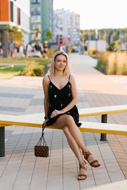 Mujer rubia alegre con un vestido negro con un bolso y gafas de sol descansa en un banco en la ciudad en verano