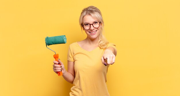 Mujer rubia apuntando a la cámara con una sonrisa satisfecha, confiada y amistosa, eligiéndote