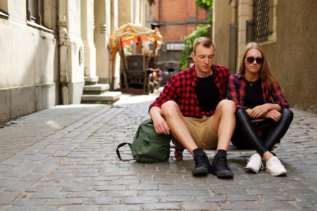 Mujer rubia atractiva con gafas de sol y su novio sentado en longboard en una calle del casco antiguo.