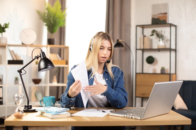 Mujer rubia con auriculares y ropa informal con video chat en una computadora portátil trabajando desde casa o en una oficina brillante