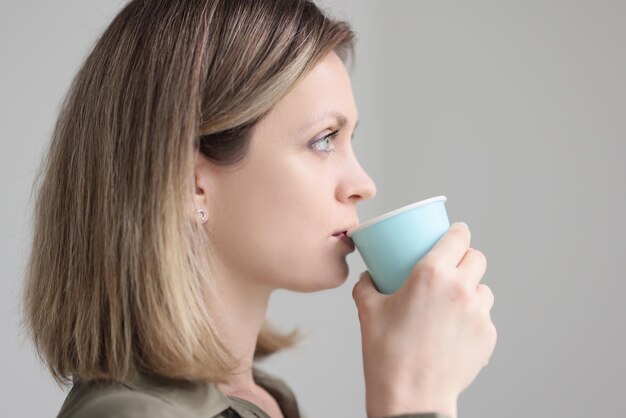 Mujer rubia bebe café de una taza de papel azul sosteniendo en la mano por la mañana sobre fondo gris