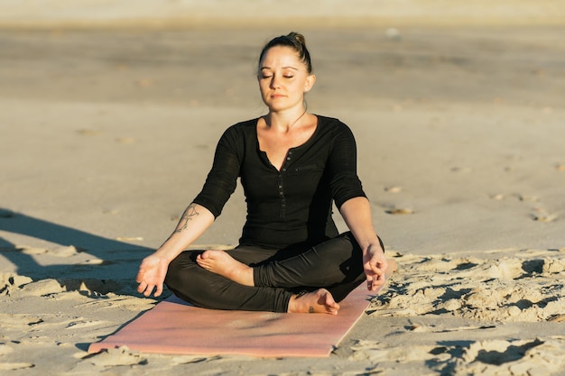 Mujer rubia de belleza caucásica en la posición de loto de yoga en una playa frente al sol