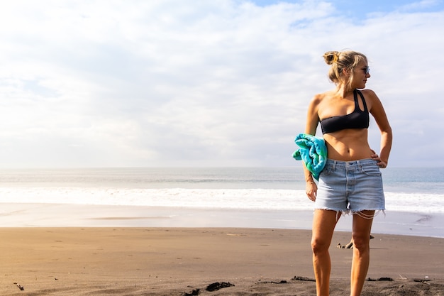 Mujer rubia en bikini y pantalones cortos de pie en una playa desierta