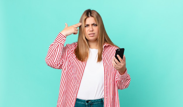 Foto mujer rubia bonita con curvas que se siente confundida y perpleja, mostrando que estás loco y sosteniendo un teléfono inteligente