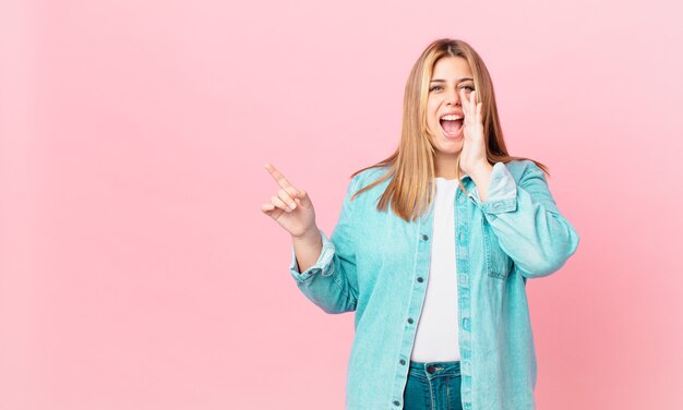 Mujer rubia bonita con curvas que se siente feliz, dando un gran grito con las manos al lado de la boca