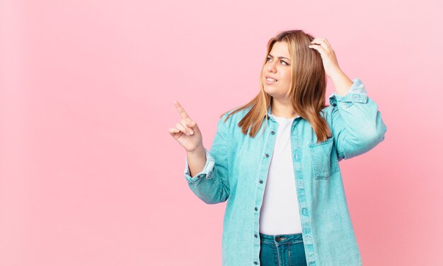 Mujer rubia bonita con curvas que se siente perpleja y confundida, rascándose la cabeza