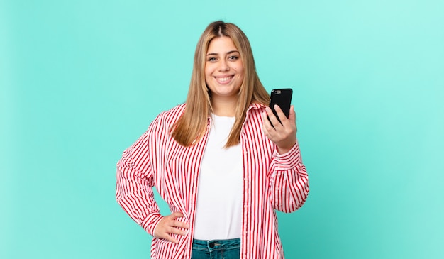 Mujer rubia bonita con curvas sonriendo felizmente con una mano en la cadera y confiada y sosteniendo un teléfono inteligente