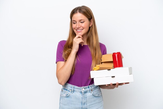 Mujer rubia bonita joven que sostiene la comida rápida aislada en el fondo blanco que mira al lado y que sonríe