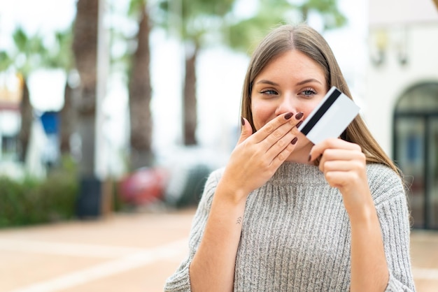Mujer rubia bonita joven que sostiene una tarjeta de crédito con expresión sorprendida