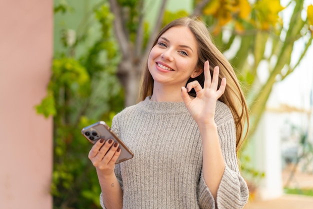 Mujer rubia bonita joven que usa el teléfono móvil al aire libre que muestra el signo de ok con los dedos