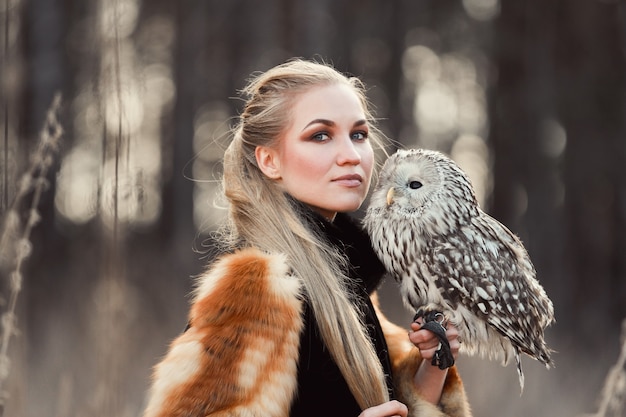 Mujer rubia con un búho en sus manos camina por el bosque en otoño y primavera. Chica de pelo largo, retrato romántico con búho. Foto de moda de arte, maquillaje hermoso