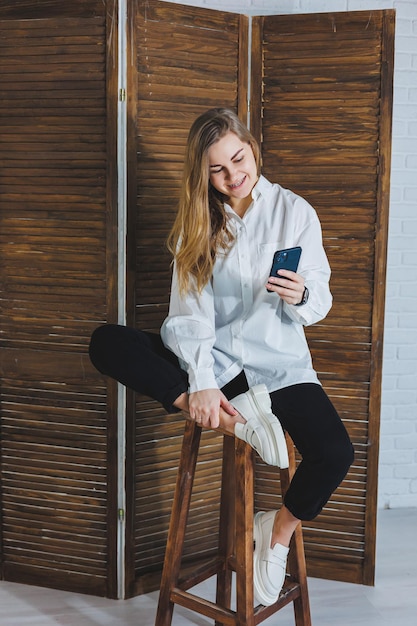 Mujer rubia con cabello largo en una camisa blanca y pantalón negro posando en la cámara del teléfono sosteniendo el teléfono y tomando un selfie en un fondo aislado