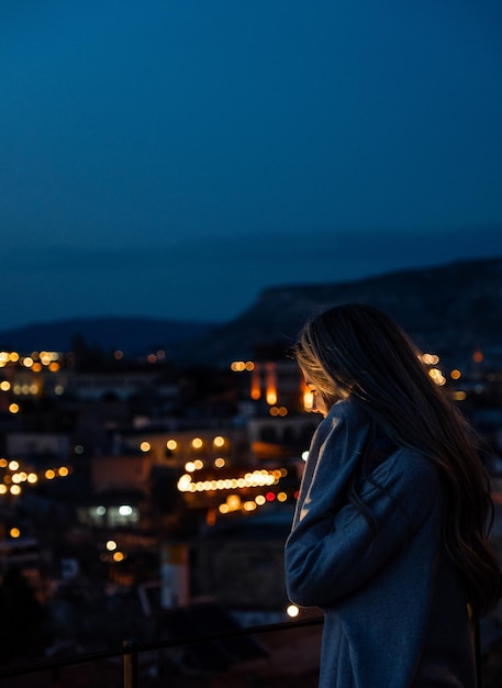 Mujer rubia con capucha mirando hacia la ciudad de Capadocia con luces de la calle por la noche