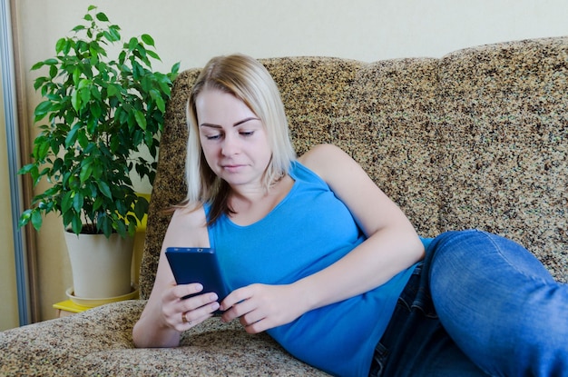 mujer rubia en casa sentada en el sofá, relájese. Trabajando en el teléfono móvil desde casa. Mujer de negocios