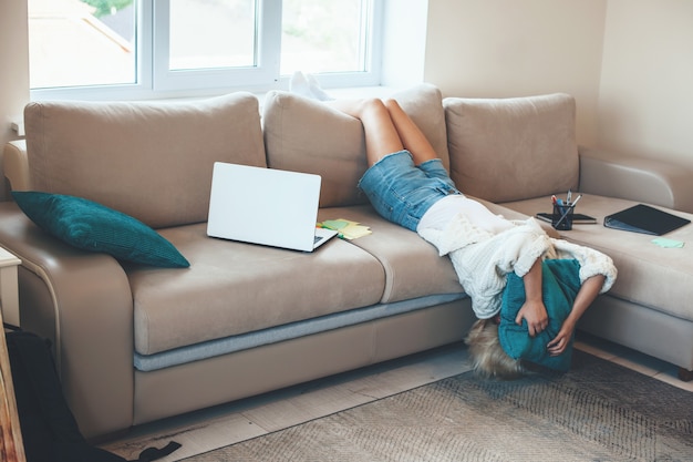 Mujer rubia caucásica agotada acostada en el sofá mientras trabaja en la computadora portátil con algunas carpetas