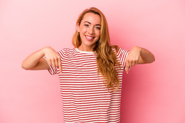 Mujer rubia caucásica aislada sobre fondo rosa apunta hacia abajo con los dedos, sentimiento positivo.