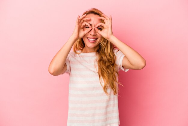 Mujer rubia caucásica aislada sobre fondo rosa mostrando bien firmar sobre los ojos