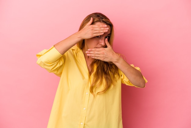 Mujer rubia caucásica aislada sobre fondo rosa parpadear a la cámara a través de los dedos, avergonzado cubriendo la cara.