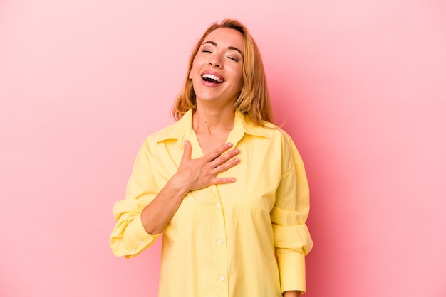 Mujer rubia caucásica aislada sobre fondo rosa se ríe a carcajadas manteniendo la mano en el pecho.