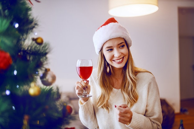 Mujer rubia caucásica alegre con el sombrero de santa en la cabeza que sonríe y que tuesta con el vino mientras que se coloca al lado del árbol de navidad en sala de estar.