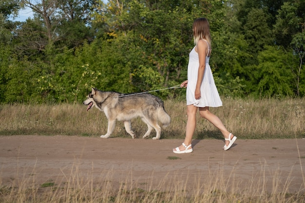 Mujer rubia caucásica en vestido blanco caminar con perro malamute de Alaska en la carretera nacional. amor y amistad entre humanos y animales.