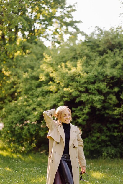 Mujer rubia caucásica wearind trinchera sonríe felizmente en un día soleado de primavera afuera caminando en el parque