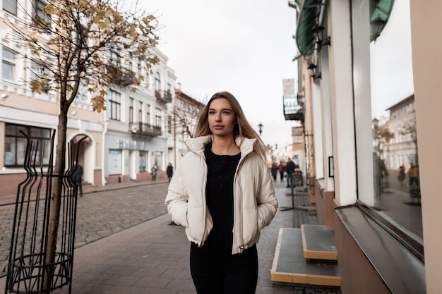 Mujer rubia con una chaqueta blanca en traje negro está posando en la ciudad