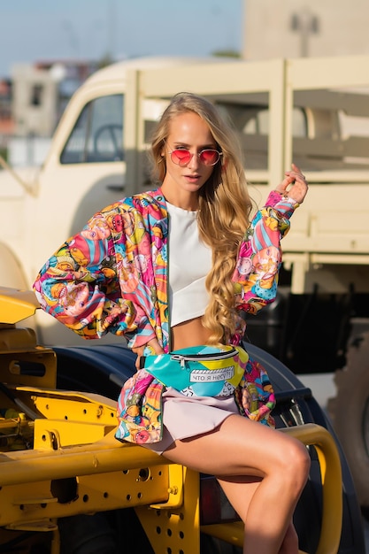 Mujer rubia con colorido traje de moda ajustando el capó y mirando a la cámara mientras se sienta en un vehículo en la calle de la ciudad