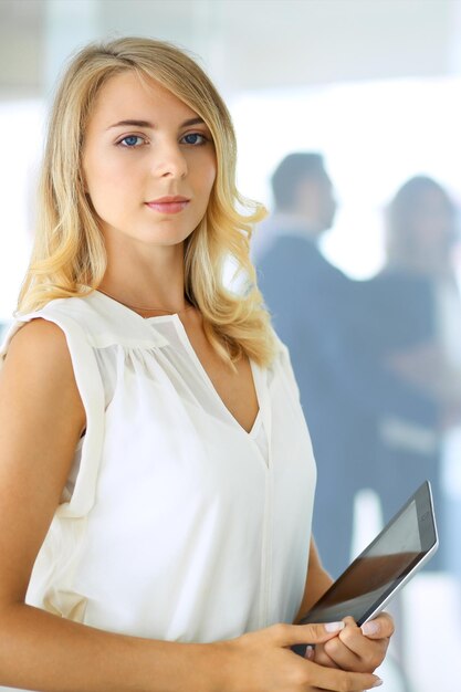 Mujer rubia con computadora touchpad mirando a la cámara y sonriendo mientras la gente de negocios se da la mano sobre el fondo