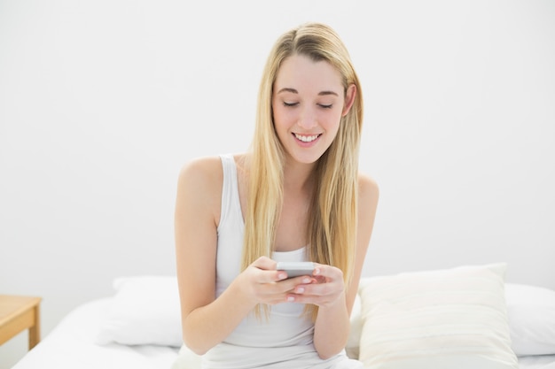 Mujer rubia contenta enviando mensajes de texto con su teléfono inteligente sentado en su cama