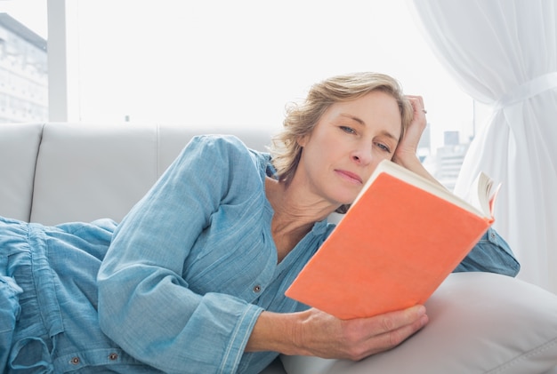 Mujer rubia contenta que se relaja en su sofá que lee el libro