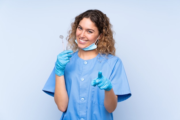 Mujer rubia dentista sosteniendo herramientas puntos aislados con el dedo hacia usted