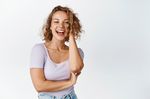 Foto mujer rubia despreocupada jugando con su cabello, riendo y sonriendo feliz, de pie en blanco. copia espacio