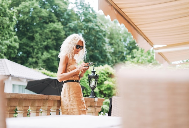 Mujer rubia elegante adulta con gafas de sol y ropa beige usando teléfono móvil en el café de la calle de la ciudad de verano