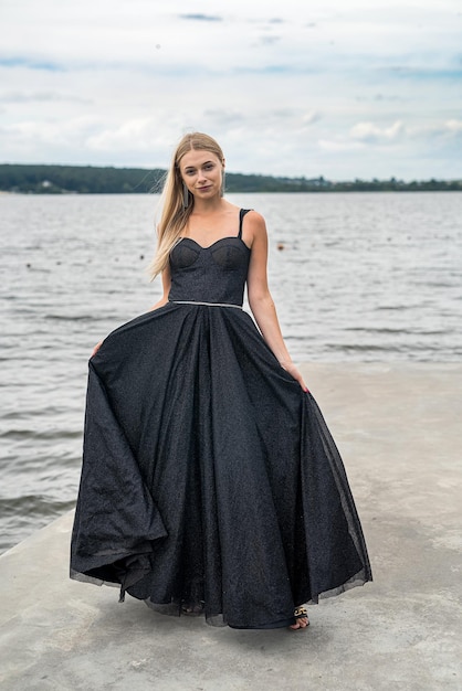 Mujer rubia elegante en un vestido de noche negro de moda cerca del lago