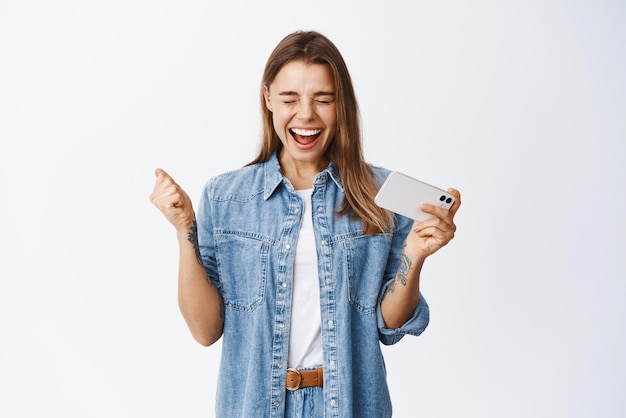 Mujer rubia emocionada gritando de felicidad sosteniendo un teléfono móvil y ganando en un videojuego en un teléfono inteligente animando y gritando fondo blanco