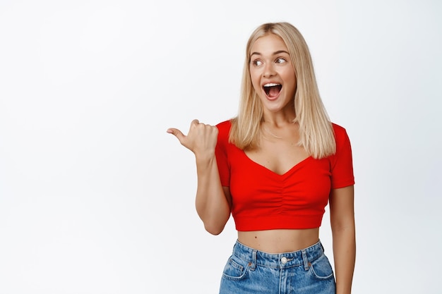 Mujer rubia emocionada señalando con el dedo y mirando a la izquierda con una expresión de asombro mirando el fondo blanco de las noticias súper geniales del anuncio