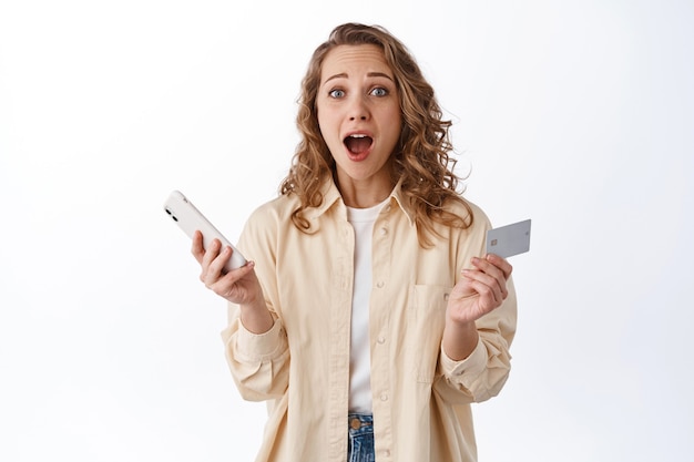 Mujer rubia emocionada y sorprendida ordena en internet, paga con teléfono inteligente y tarjeta de crédito, jadeando en el frente, pared blanca