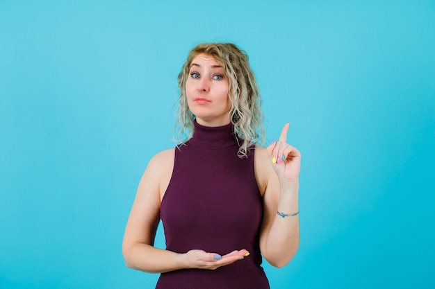 Mujer rubia está apuntando hacia arriba con el dedo índice sobre fondo azul.