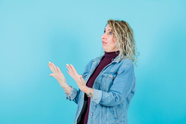 Mujer rubia está mirando hacia arriba y levantando las manos sobre fondo azul.