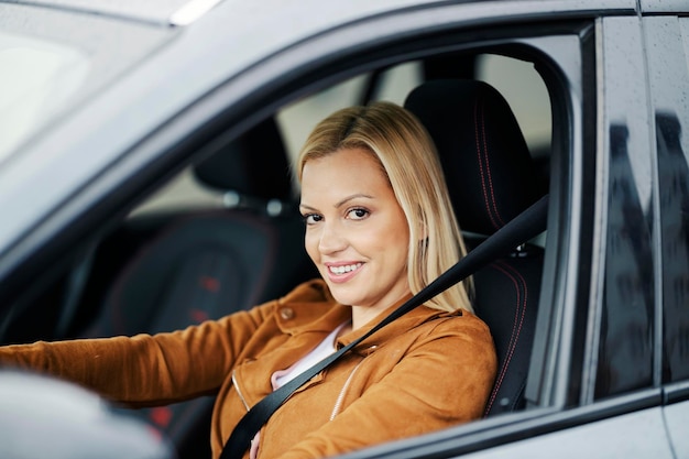 Una mujer rubia feliz conduce su auto mientras sonríe a la cámara